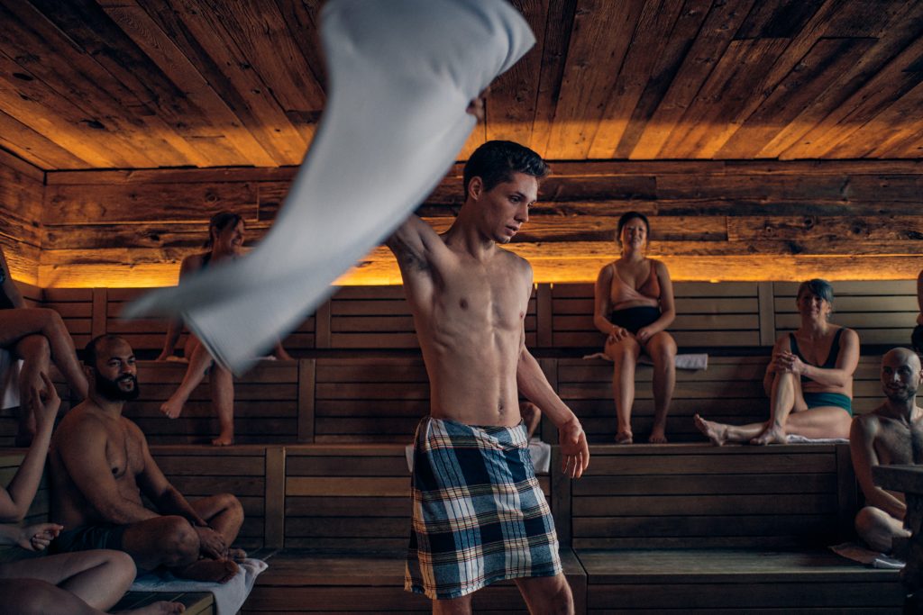 A Ritual Artisan energetically waves towels during his Aufguss Ritual. The artisan stands amidst billowing clouds of steam, their movements fluid and precise. The towels create mesmerizing patterns as they slice through the air, enhancing the sensory experience of the ritual.